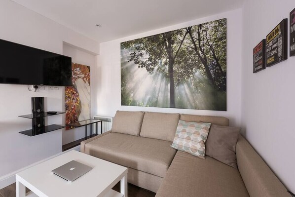 A modern living room with a beige sofa, a flat-screen TV mounted on the wall, and a large forest-themed painting above the sofa. - Spectacular Modern Apartment (5 mins to tube) (Greater London)