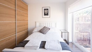 A modern bedroom with a wooden wardrobe, a bed with white linens, a framed artwork on the wall, and a view through a large window. - Spectacular Modern Apartment (5 mins to tube) (Greater London)