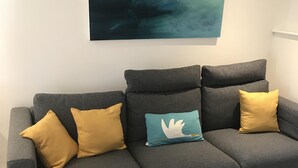 Een grijze bank met mosterdkleurige en smaragdgroene kussens, een klein rond tafeltje en een blauw pluisachtig vloerkleed op een lichtgekleurde vloer.