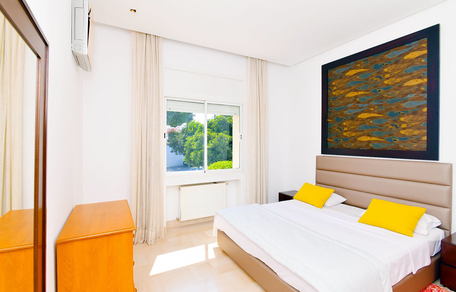 A hotel room with a bed, a window with curtains, a radiator, and a painting on the wall.