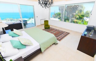 Une chambre à coucher comprenant un lit, un bureau, une chaise, un lustre et de grandes fenêtres donnant sur la mer.