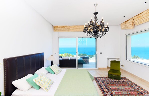 A bedroom with a bed, a green armchair, a chandelier, and a view of the sea through a sliding door.