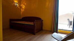 A room with a wooden floor, a bed with a rainbow-striped blanket, a wall-mounted light fixture, and a view of an outdoor balcony with a chair. - La Villa Sanary-Sur-Mer (Sanary-sur-Mer)