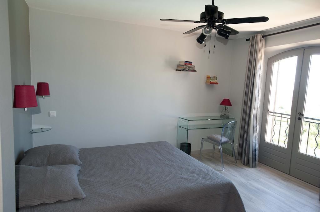A bedroom with a bed, a desk, and a chair. There is a ceiling fan, a window with curtains, and a balcony.