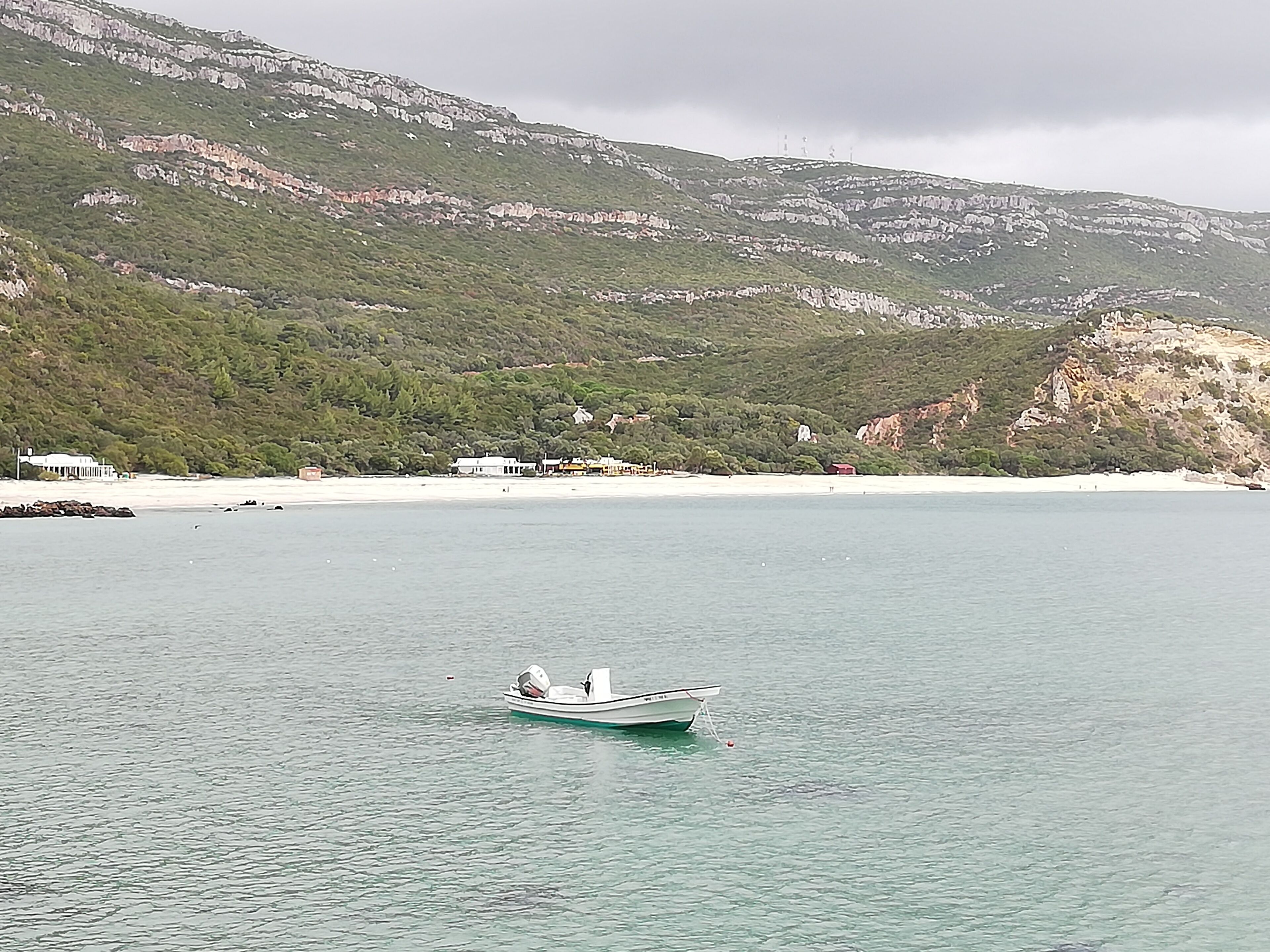 Un bateau sur une étendue d’eau calme, près d’une zone côtière, avec des bâtiments et une chaîne de montagnes en arrière-plan.