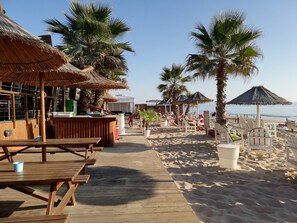 A beachside restaurant with outdoor seating, palm trees, and a wooden deck leading to the beach. - Casa da Vista Rio (Amora)