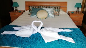 A neatly made bed with a heart-shaped towel arrangement, a blue textured bedspread, and two bedside lamps on wooden nightstands. - Casa da Vista Rio  (Amora)