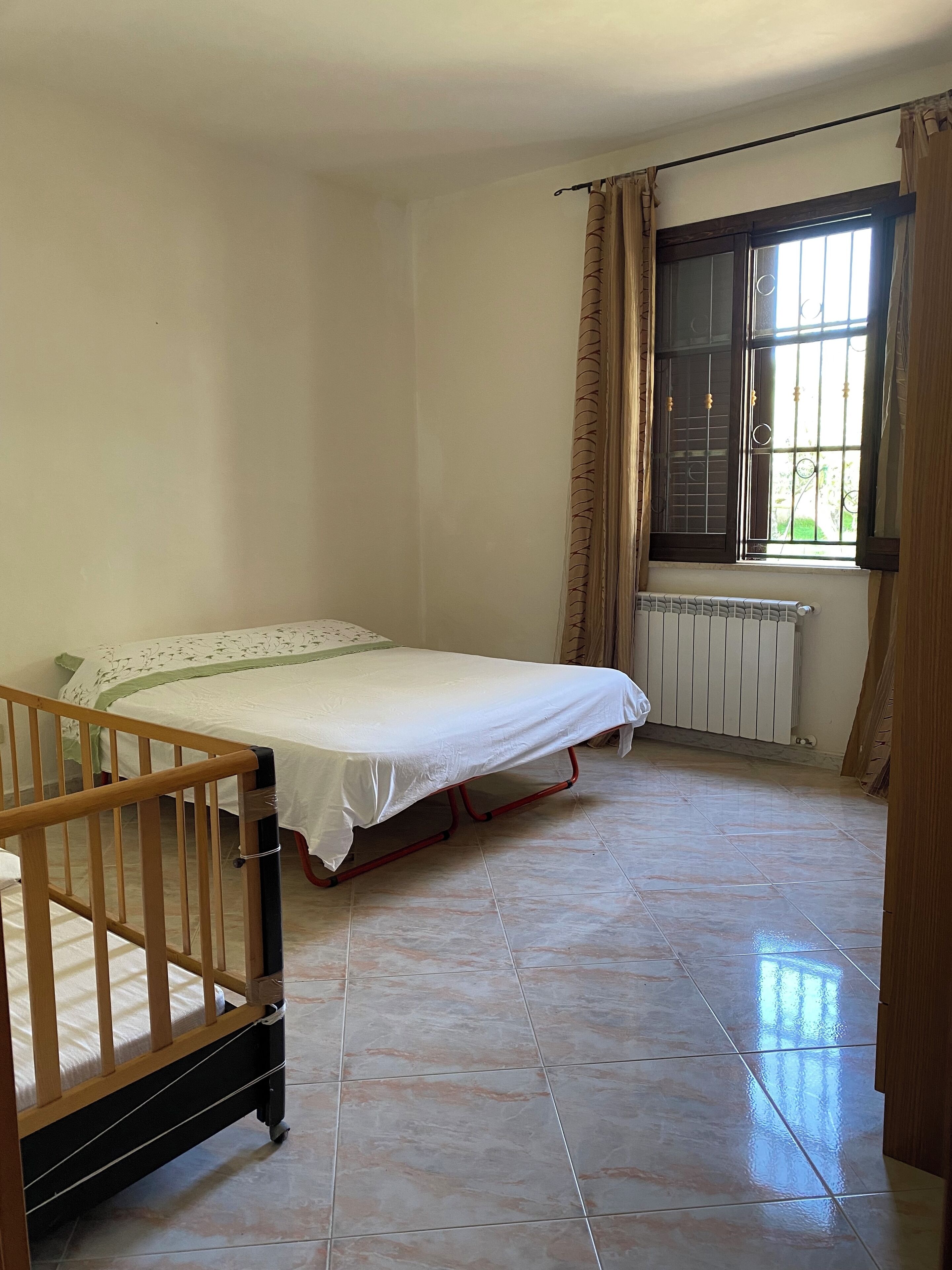 Une chambre avec un lit simple, un berceau en bois, un radiateur et une fenêtre avec des rideaux.