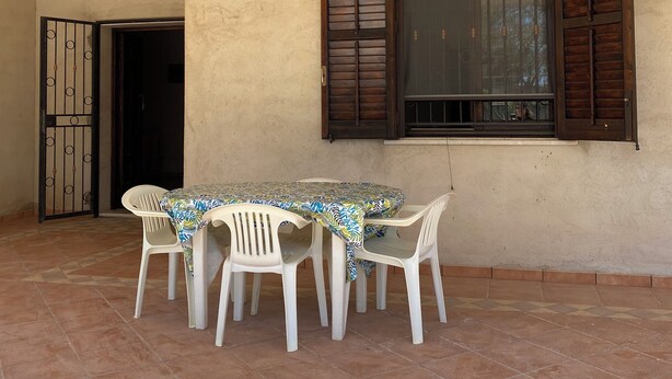 Eine überdachte Terrasse mit Tisch und Stühlen, ein Fenster mit Rollläden sowie eine Tür mit einem Metallgitter.