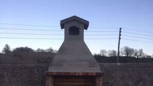 Een bakoven van baksteen met schoorsteen, geplaatst op een betonnen basis, met op de achtergrond een glooiend landschap en elektriciteitskabels.