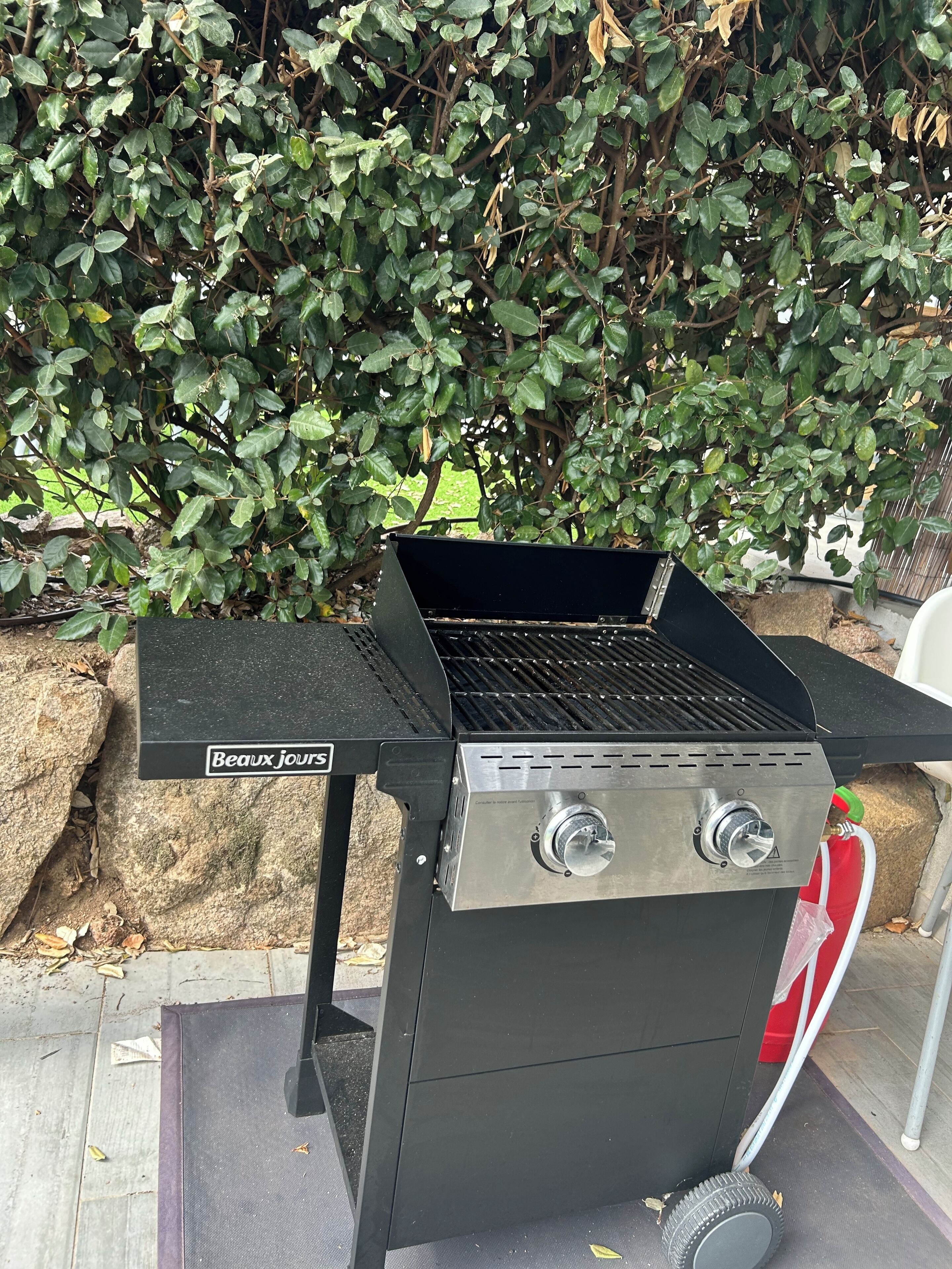 A black barbecue grill with two control knobs and a side burner, labeled "Beaux Jours," set against a backdrop of dense green foliage.