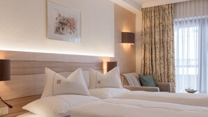 Una habitación de hotel con una cama grande, una mesita de noche, una lámpara de pared, una obra de arte floral enmarcada, un sofá con cojines y una ventana con cortinas.