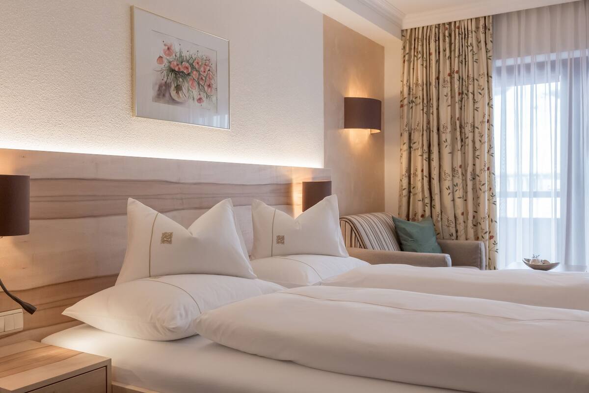 Hotellihuone, jossa on suuri sänky, yöpöytä, seinään kiinnitetty lamppu, kehystetty kukka-aiheinen taideteos, tyynyillä varustettu sohva ja ikkuna, jossa on verhot.