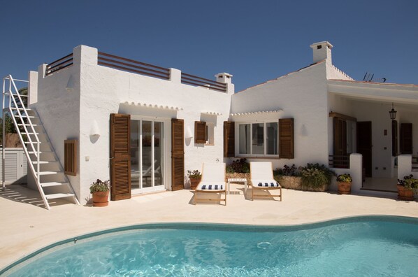 A white villa with a pool, wooden shutters, and a staircase leading to the roof.