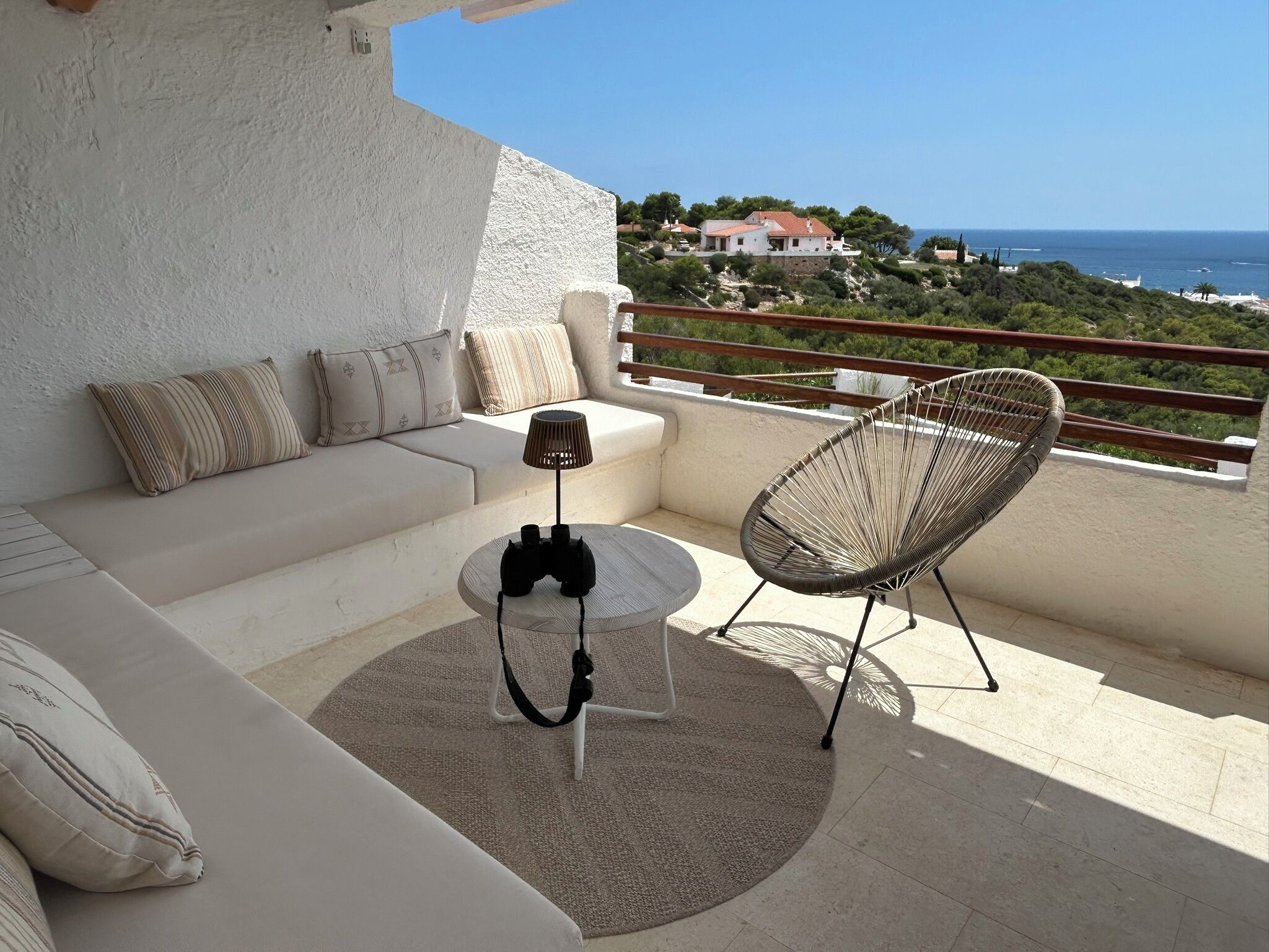 A balcony with a sofa, a chair, a small table, and a lamp. The balcony overlooks a scenic view of the sea and a distant building.