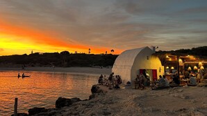 A beachside restaurant with a white dome structure, outdoor seating, and a view of the sunset over the water.