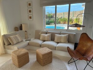 A living room with a beige sectional sofa, two woven ottomans, a brown leather chair, and a view of a pool and garden through large windows.
