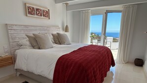 A bedroom with a bed, a red blanket, a patterned rug, a wooden headboard, and a view of the sea through the window.
