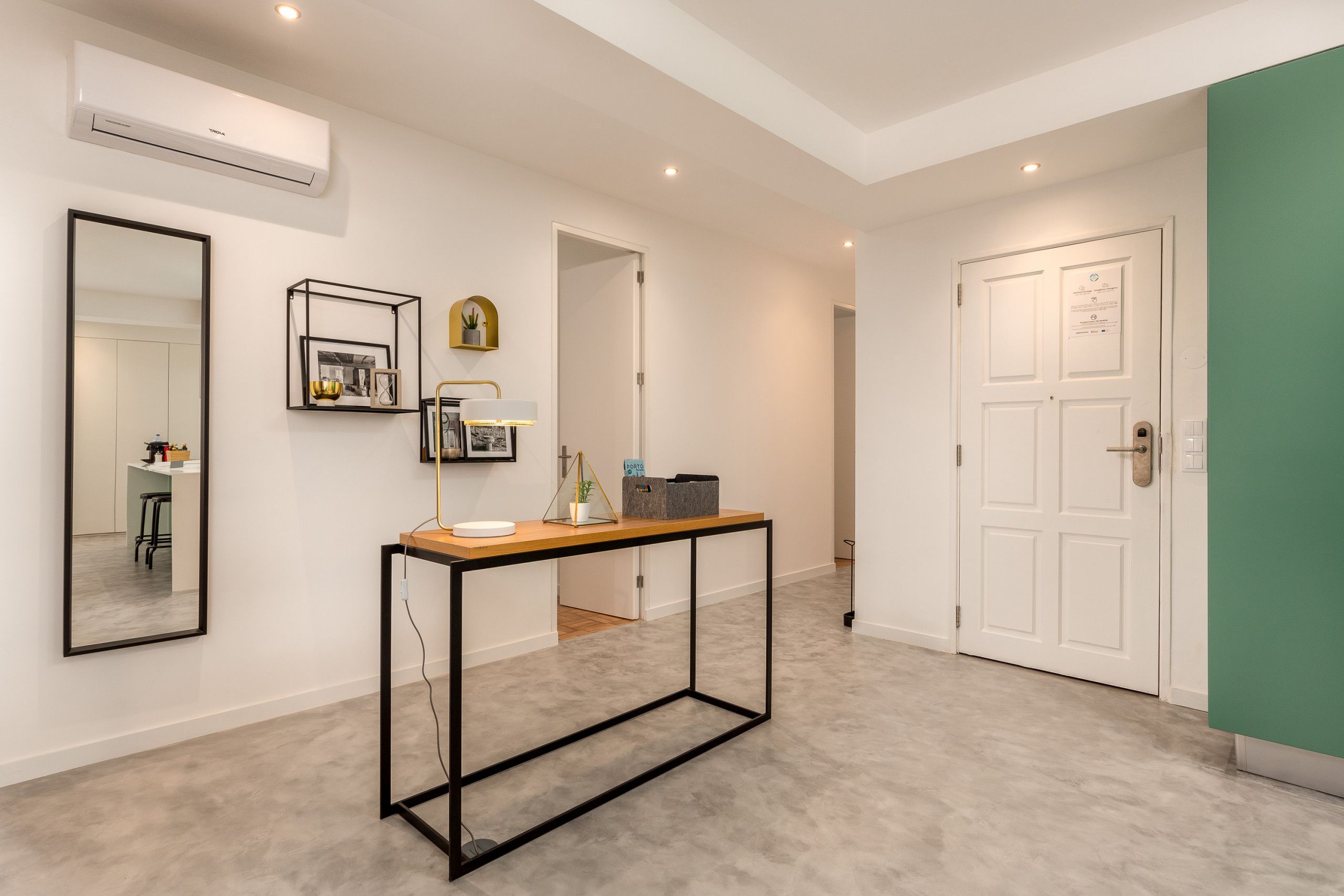 Een moderne hal met een minimalistisch ontwerp, met een console tafel, een lamp, een lijst met een afbeelding en een kleine plant. Aan de rechterkant een witte deur met een plaquette en een groene muur.