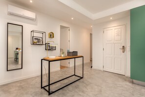 A modern hallway with a minimalist design, featuring a console table with a lamp, a framed picture, and a small plant. There is a white door with a plaque and a green wall on the right.