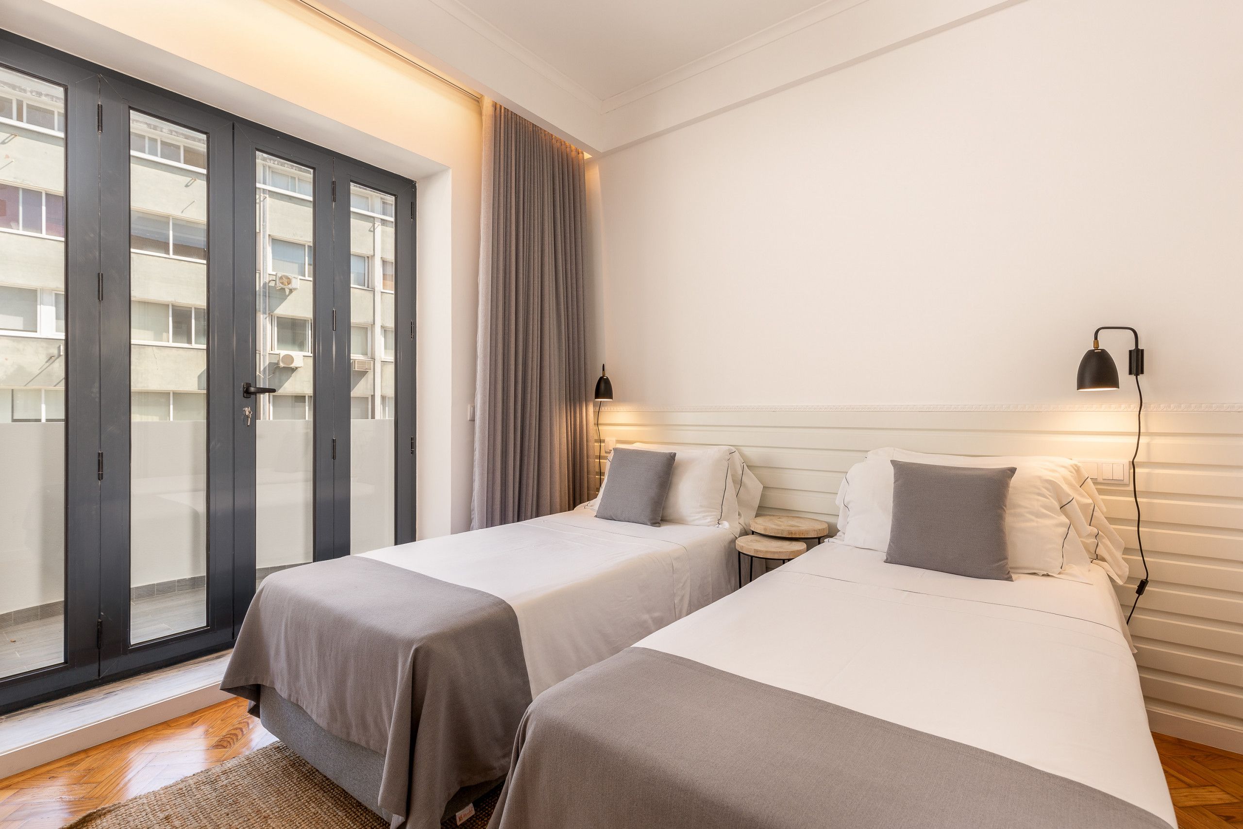 Habitación de hotel con dos camas, una mesita de noche pequeña, una lámpara y vistas a través de una puerta corrediza.