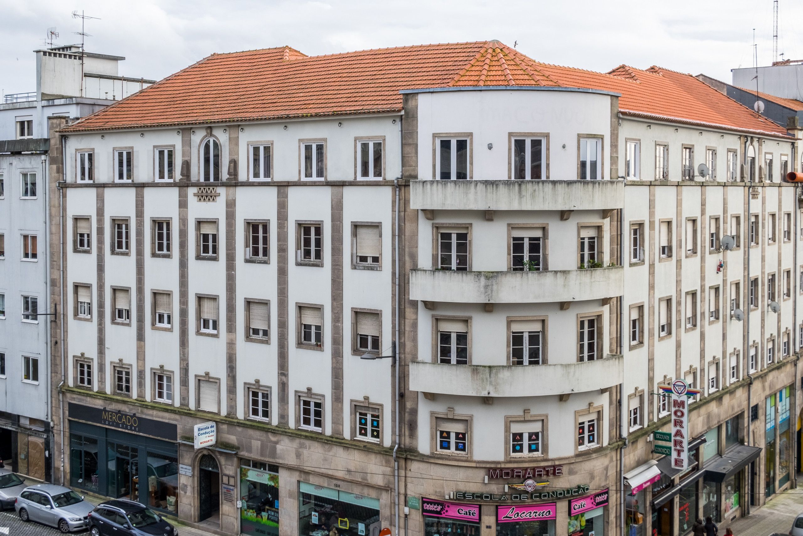 Ein mehrgeschossiges Gebäude mit rotem Ziegeldach und weißer Fassade, mit Geschäften im Erdgeschoss und einem gut sichtbaren Eingang.