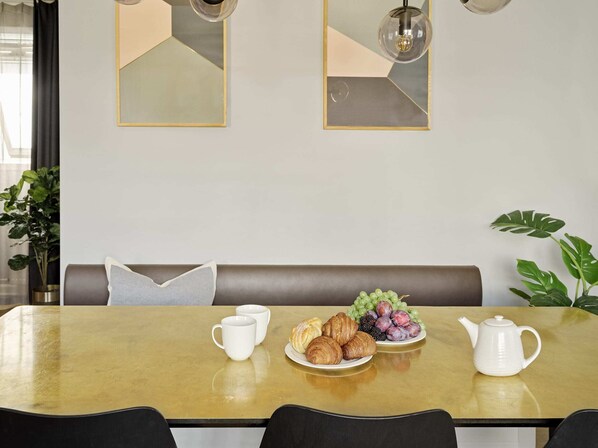 A dining area with a table set for two, featuring a plate of croissants, grapes, and blackberries, a white teapot, and two white cups.