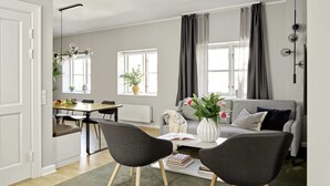A modern living room with a grey sofa, two black chairs, a white coffee table with books, a vase with flowers, and a dining area with a table and chairs. - Spacious Two-Bedroom Apartment for 6 with Elevator (København)