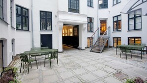 Une cour intérieure avec un escalier menant à l’étage supérieur, un espace extérieur aménagé avec des tables et des chaises, et une entrée de bâtiment avec une porte noire.
