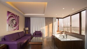 A modern hotel room with a purple sofa, a glass coffee table, a large window with a city view, and a separate bathroom with a bathtub. - The Opera Hotel (Kfar Yassine)