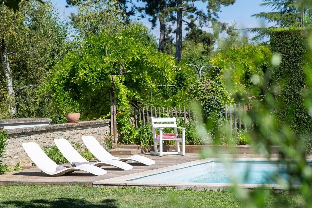 Zona de piscina con sillones de descanso y una mesa pequeña, rodeada de follaje y árboles.