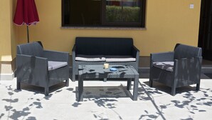 Un área de estar al aire libre con dos sillas de mimbre y una mesa, una sombrilla roja y una ventana con persianas.