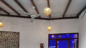 An indoor dining area with white plastic chairs and red tablecloths, a ceiling fan, hanging light fixtures, and a stone wall decoration. - Alkina Lodge (Fulidhoo)