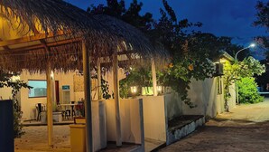 A thatched-roof outdoor seating area with a concrete bench and a small table, surrounded by greenery and a building with a stone wall. - Alkina Lodge (Fulidhoo)