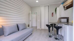 A modern living room with a sofa and two bar stools, a kitchen area with a microwave, and a flat-screen TV on the wall.