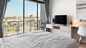 A modern hotel room with a large bed, a TV on a white console table, a desk with a chair, and a balcony with a view of the city. - Nidhra Residence (Lam Luk Ka)