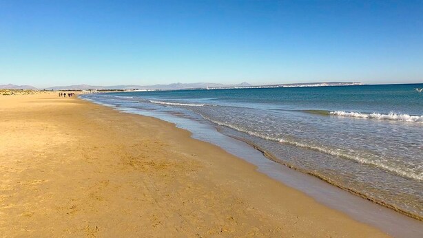A wide sandy beach with gentle waves, a clear sky, and distant mountains.