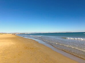 Une vaste plage de sable fin, avec des vagues douces, un ciel dégagé et des montagnes à l’horizon.