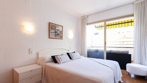 A bedroom with a bed, a nightstand, a wall-mounted light, a painting, and a view through a sliding door. - Nice family apartment close to PortAventura - UHC JEREZ APARTMENTS 158 (Salou)