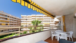 A balcony with white plastic chairs and a table, a view of a building with multiple balconies, and a palm tree in the background. - Nice family apartment close to PortAventura - UHC JEREZ APARTMENTS 158 (Salou)