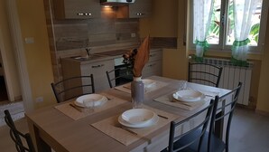 Un comedor con una mesa dispuesta para cuatro personas, incluyendo platos, vasos y cubiertos. Hay un florero con una planta seca sobre la mesa. En el fondo se ve una cocina con salpicadero de azulejos y armarios de madera.