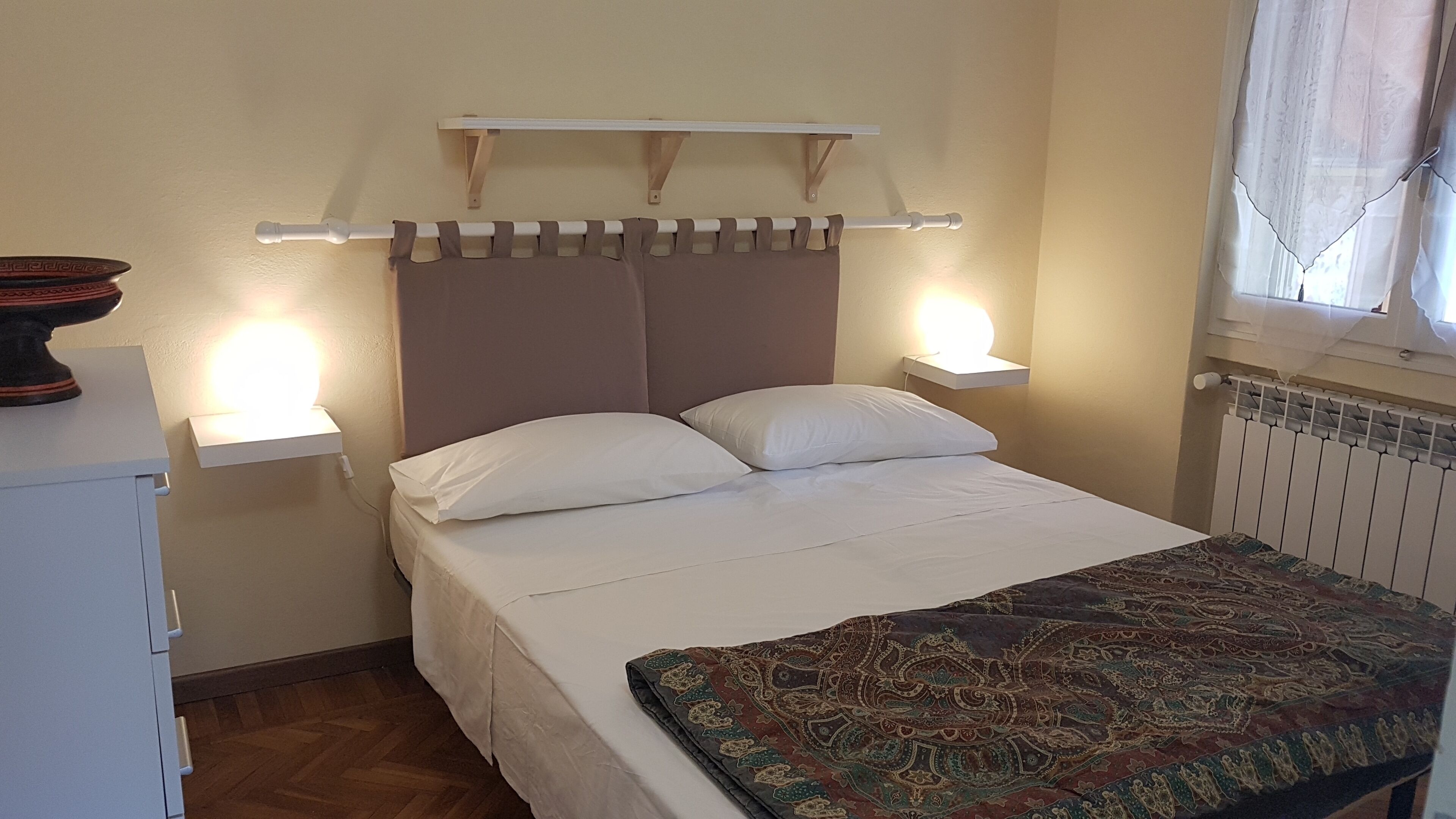 Een slaapkamer met een bed, nachtlampjes, een nachtkastje, een radiator en een raam met gordijnen.