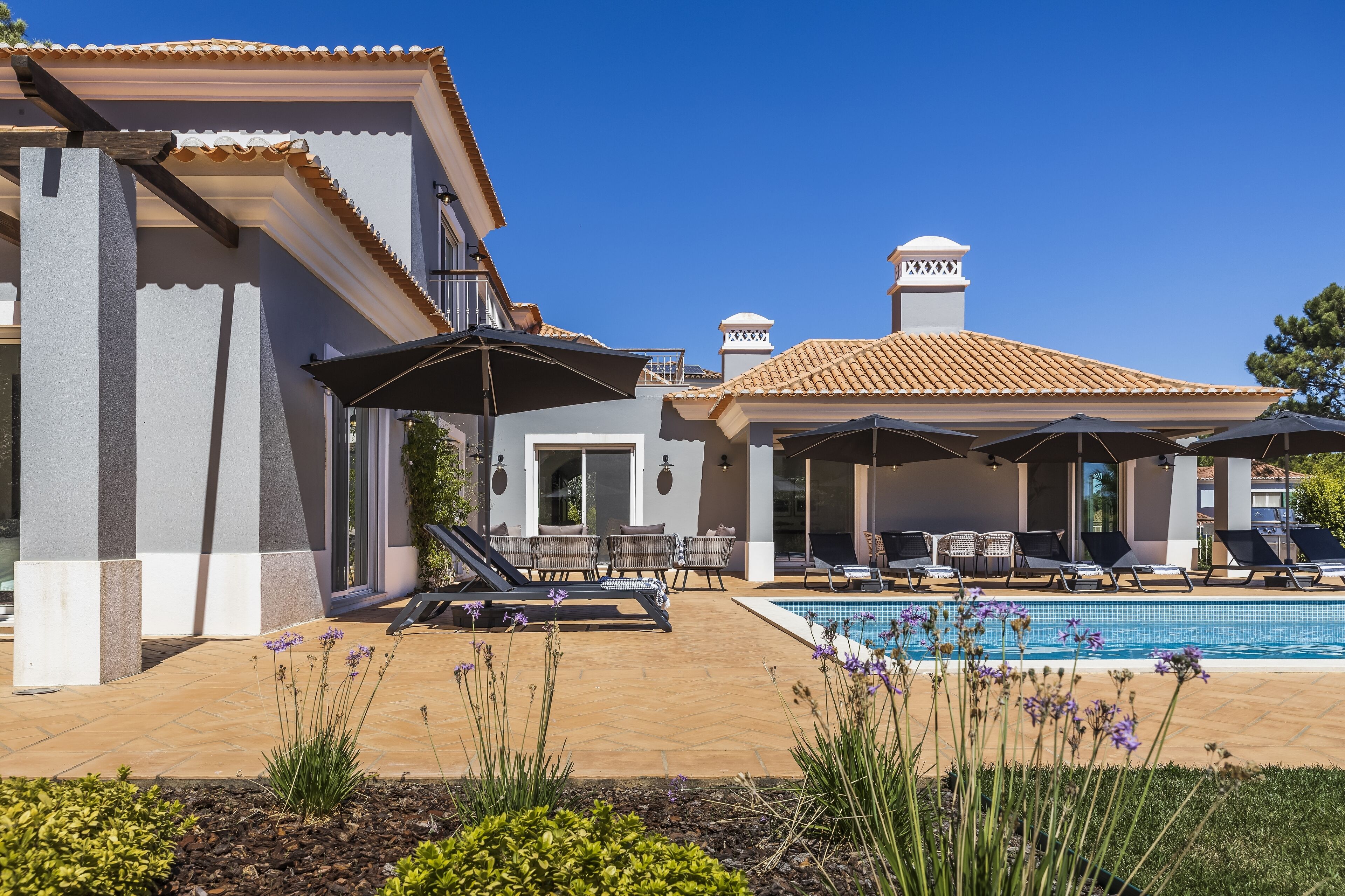 A poolside area with lounge chairs and umbrellas, a swimming pool, and a building with a tiled roof and columns.