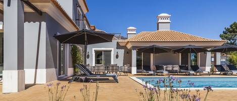 A poolside area with lounge chairs and umbrellas, a swimming pool, and a building with a tiled roof and columns.