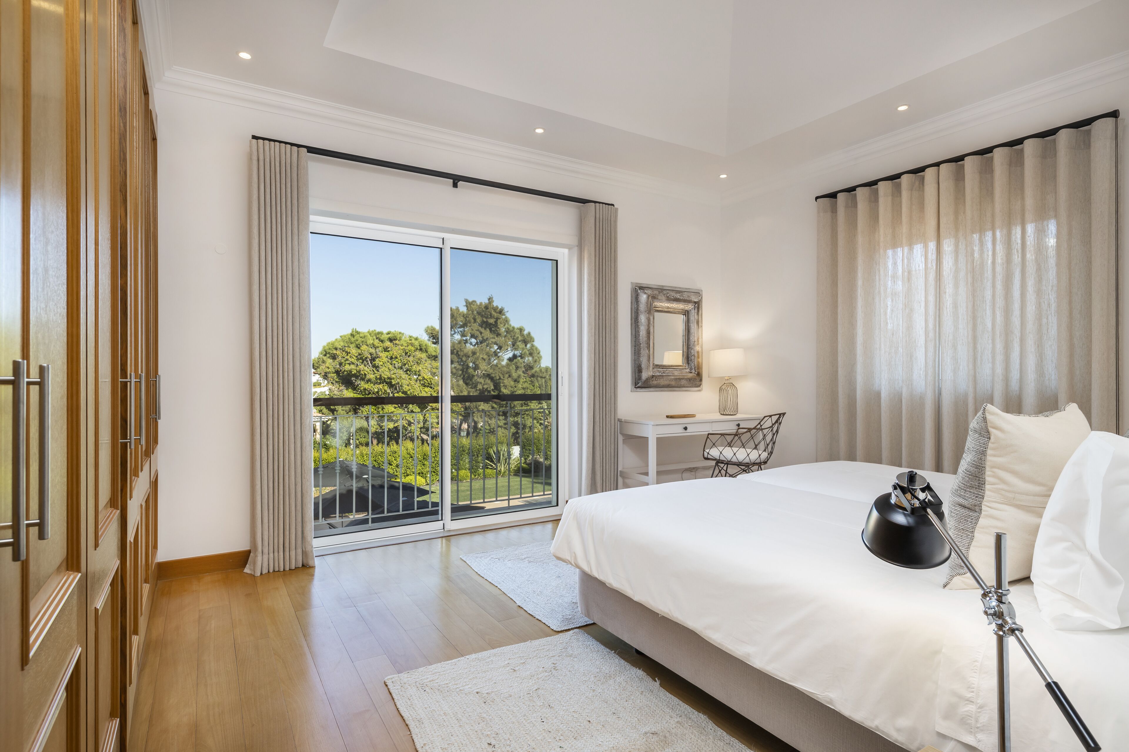 A bedroom with a large bed, a desk, and a view of greenery through a sliding door.