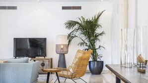 Ein modernes Wohnzimmer mit einer Couch, einem Korbsessel, einem Fernseher, einer Stehlampe, einer großen Kübelpflanze und dekorativen Glasvasen.