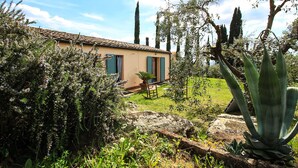 A rustic house with a tiled roof, surrounded by lush greenery and tall cypress trees.