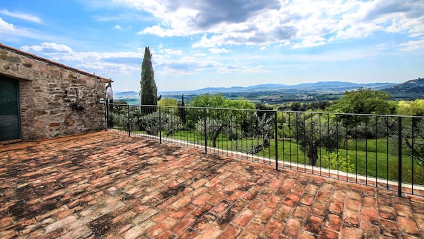 A terrace with a stone wall, a metal fence, and a view of a vineyard and distant hills.