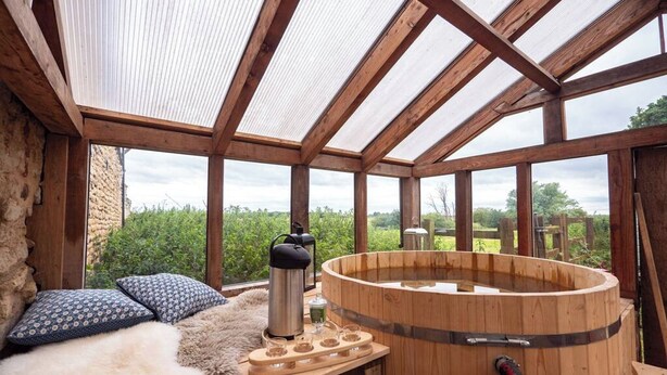 A wooden room with a hot tub, a bed with pillows, a fur throw, and a table with glasses and a thermos.