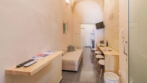 A narrow hallway with a wooden bench, a sofa, and a table with chairs. A TV is mounted on the wall, and a door is visible on the right. - Balbo 9A Apartment - Lecce Selection (Lecce)